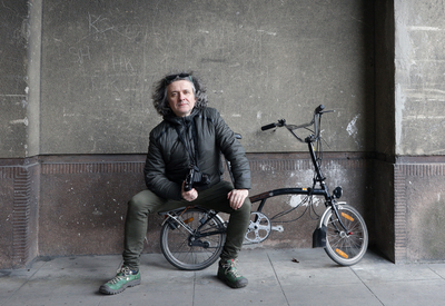 Maciej Rawluk, a middle-aged man with shoulder-length gray hair, sitting on a foldable bike with a camera in his hand 