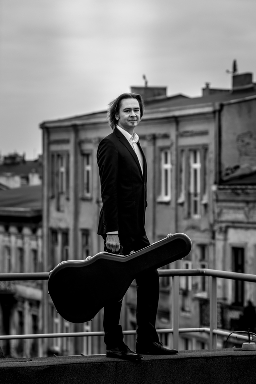 Maciej Staszewski wearing a suit and standing with a guitar on the balcony, with tenement houses visible in the background